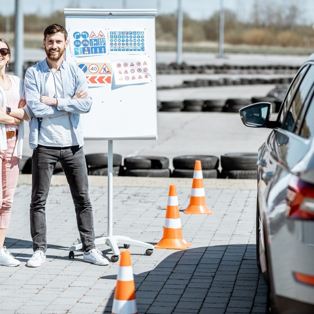 Portrait of an instructor and female student standing with road signs and learning car on the training ground at the driver's school